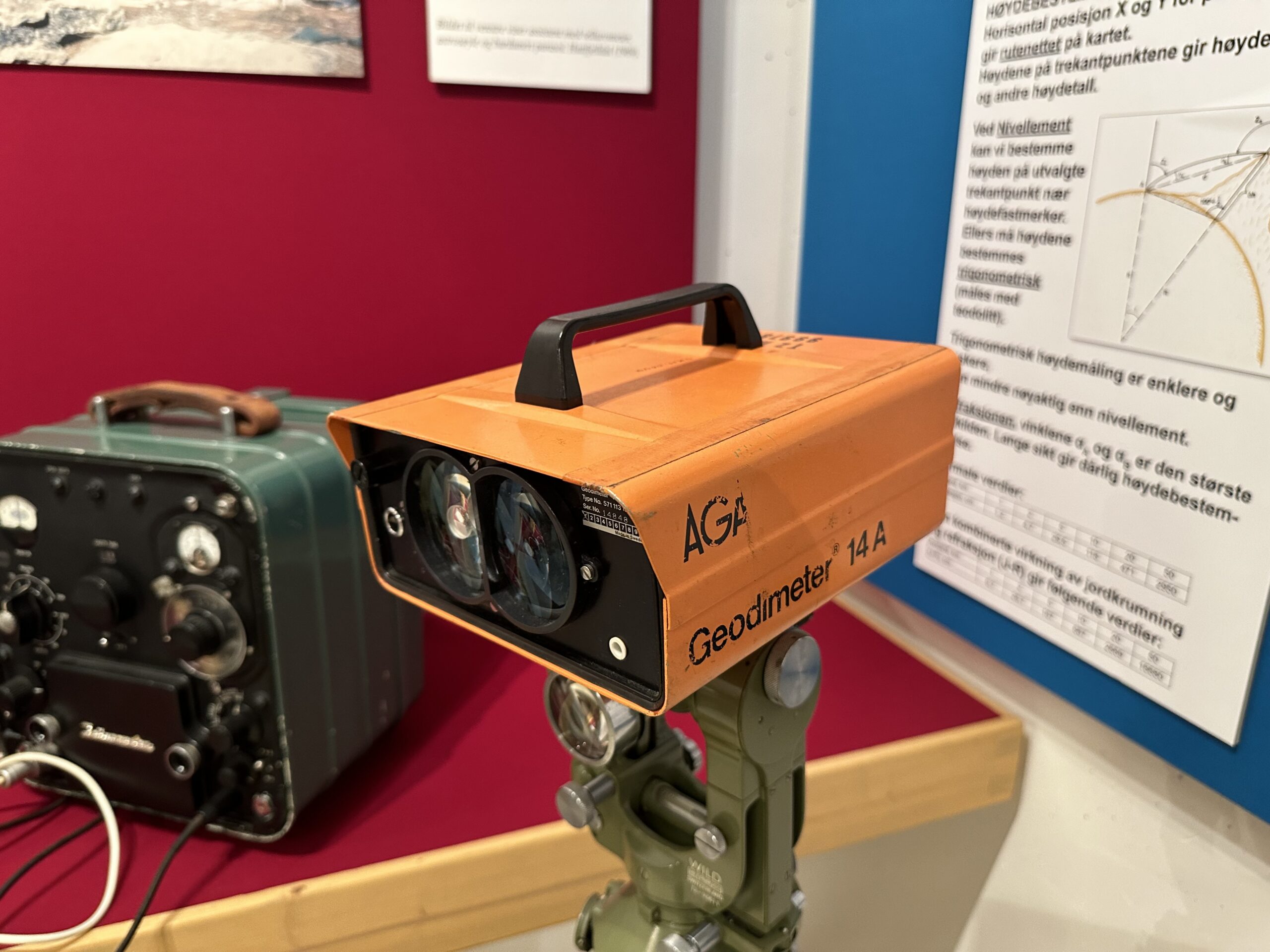 An orange AGA Geodimeter 14A sits on a tripod in a museum display, with other surveying equipment beside it.