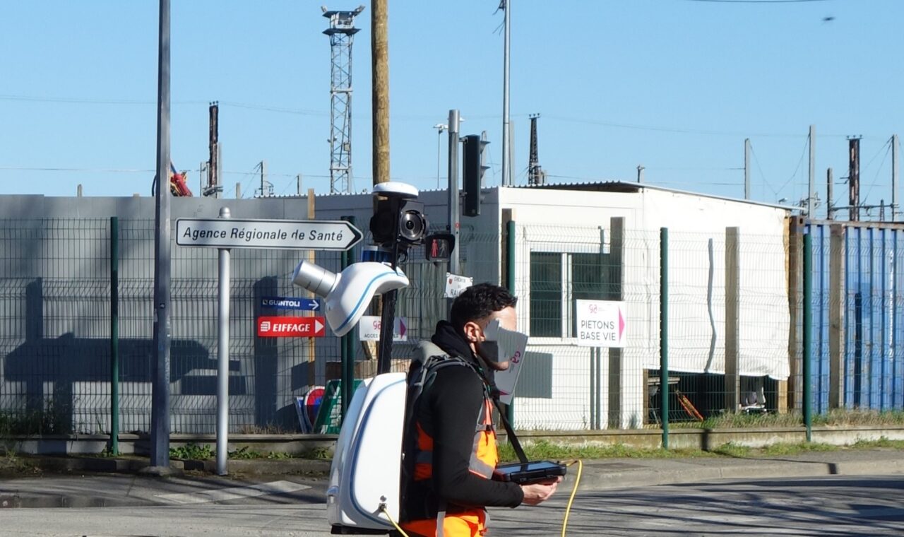 A person walks along a street wearing a tall backpack-mounted mapping sensor assembly.