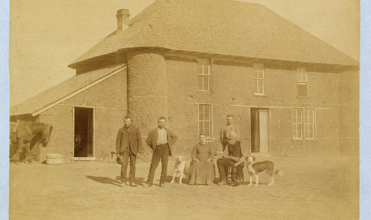 The Haumont family and dogs posed in front of a two-story sod house with a broad shingled roof in Nebraska in 1886.