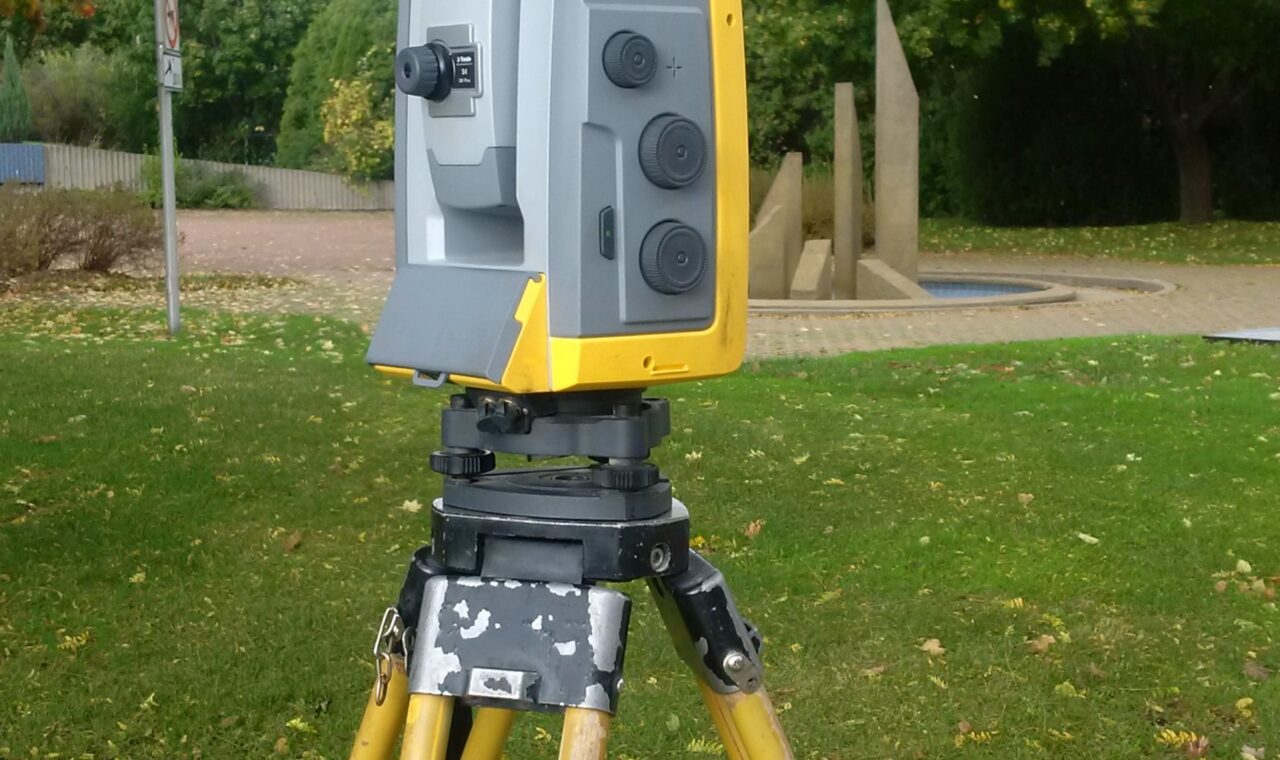 A yellow and gray robotic total station stands on a yellow tripod in a grassy setting.