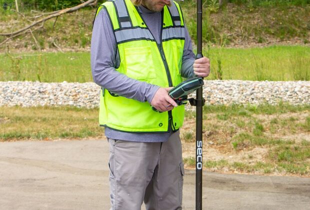 A surveyor holds a GNSS pole and controller over a painted parking stripe, with trees in the background.