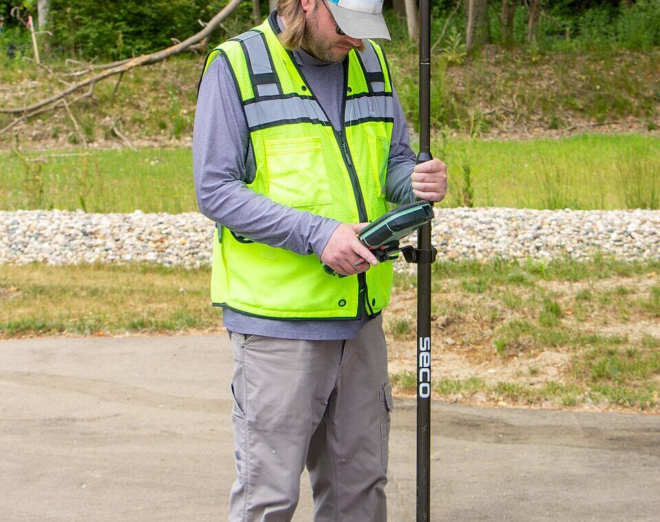 A surveyor holds a GNSS pole and controller over a painted parking stripe, with trees in the background.
