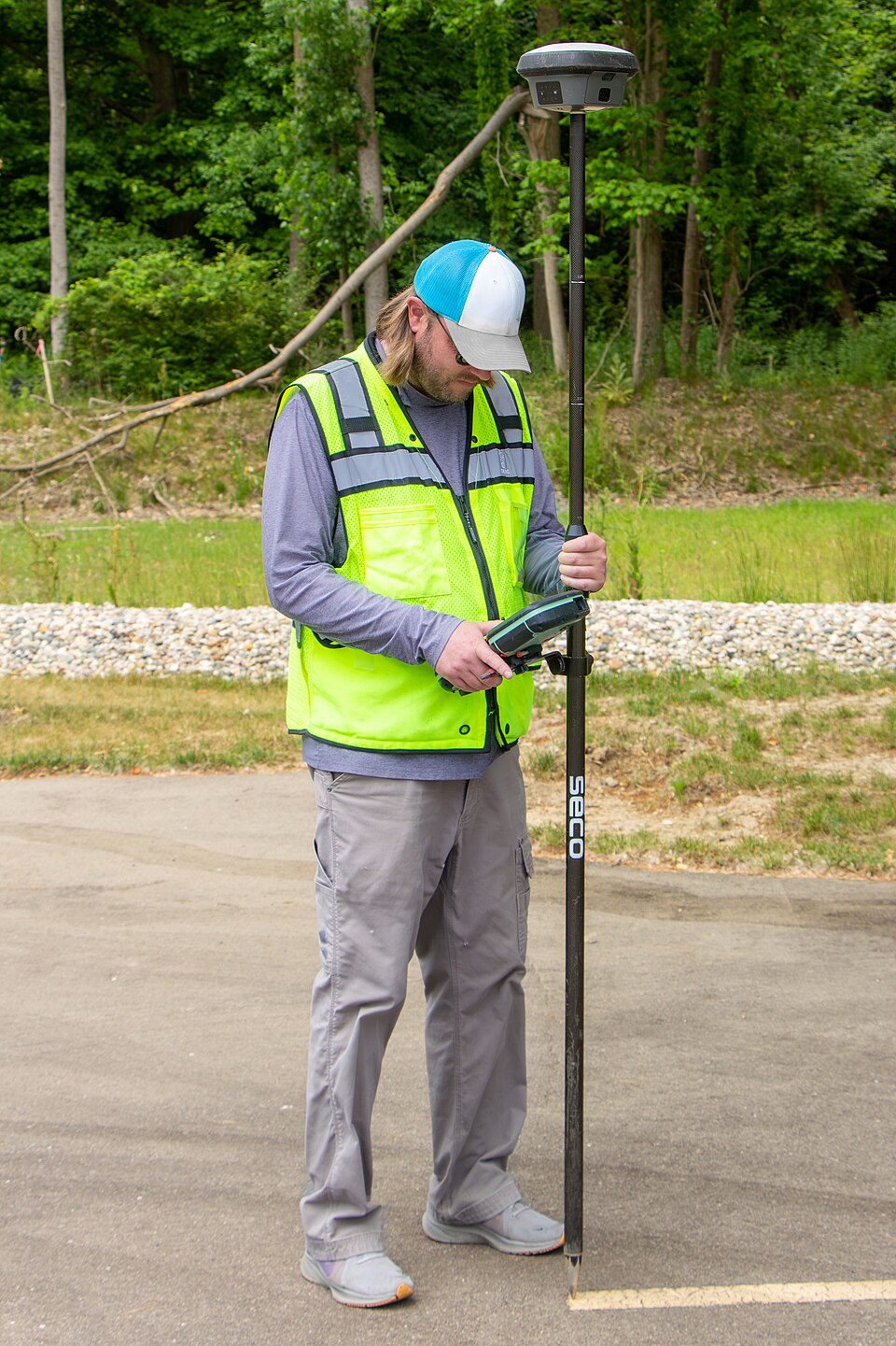 A surveyor holds a GNSS pole and controller over a painted parking stripe, with trees behind him.