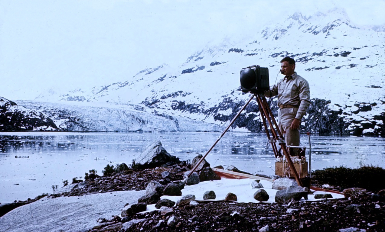 A surveyor stands beside a tripod-mounted Tellurometer on a rocky shore overlooking Columbia Glacier in Alaska.