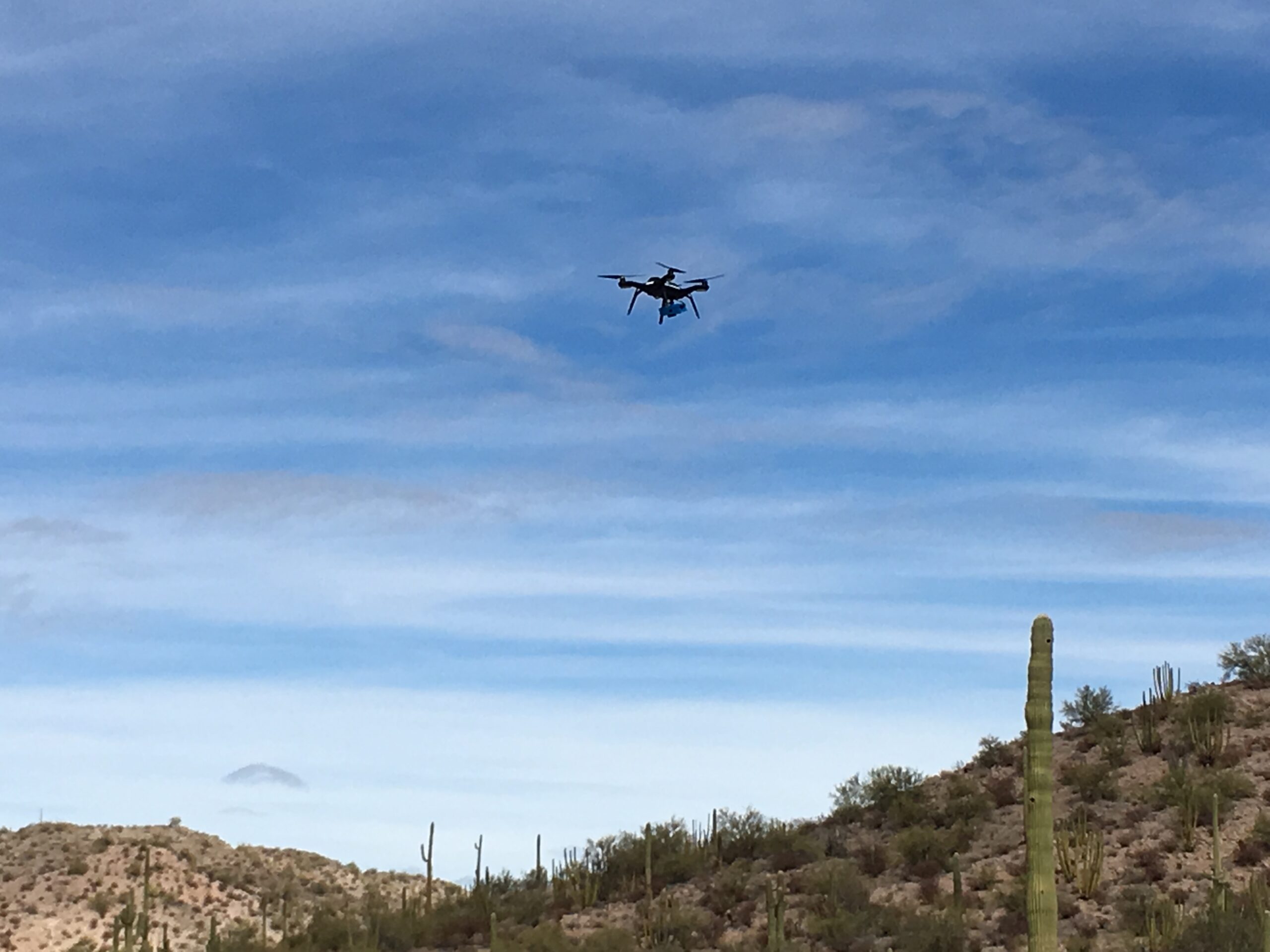 A small quadcopter flies over a desert landscape with saguaro cacti beneath a blue sky.